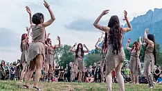 Gruppo di danzatori disposti in cerchio in un'azione coreografica all'aperto, con movimenti delle braccia sincronizzati. Indossano costumi naturali ispirati a tessuti grezzi, immersi in un paesaggio montano con pubblico sullo sfondo. Scena del laboratorio coreografico Fairy Circle dedicato alla danza rituale e alla connessione con il movimento collettivo