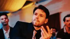 Un primo piano di Marcello Merighi in un contesto con luci soffuse e calde, probabilmente una sala da ballo. Indossa un elegante abito scuro, una camicia nera e una cravatta a fantasia sui toni dell'oro e del marrone. È ritratto in una posa dinamica, con il busto leggermente inclinato e le mani sollevate e aperte in un gesto tipico della danza, come se stesse eseguendo un passo di Lindy Hop o Swing. Il suo sguardo è intenso e rivolto in alto a destra. Sullo sfondo, fuori fuoco, si intravedono altre persone.